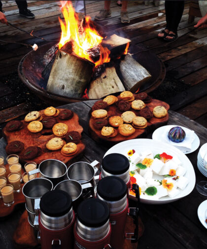 Beside a roaring campfire is a table holding food and drinks.