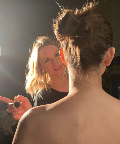 A makeup artist faces the camera as she works on a model, who is facing away from us.