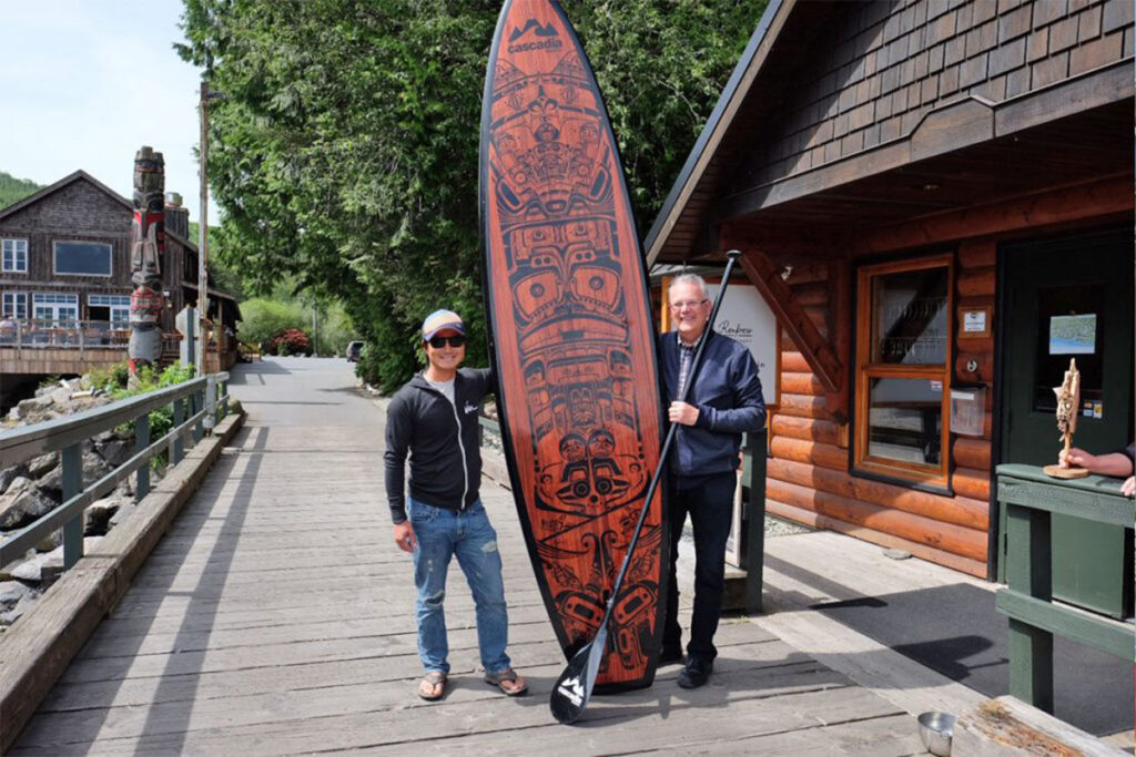 Two men stand beside a surfboard. 