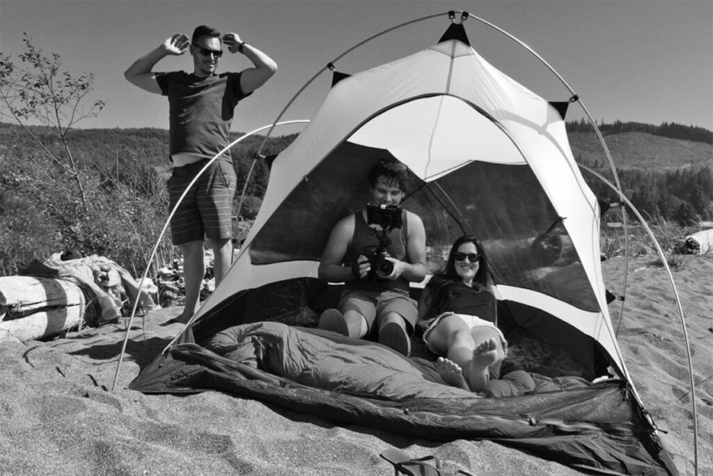 Two people lounge in a tent on the sand, while a man stands nearby, stretching. 