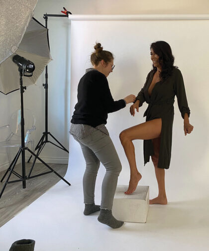 A woman adjusts a model's sleeve for a photoshoot. Both are smiling.